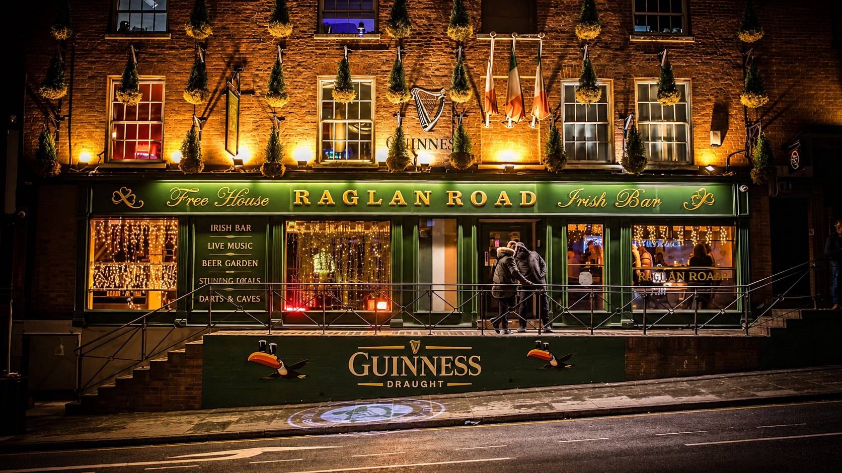 The Raglan Road Irish Bar shopfront on Derby Road at night, lit gold and green above the Guinness wall